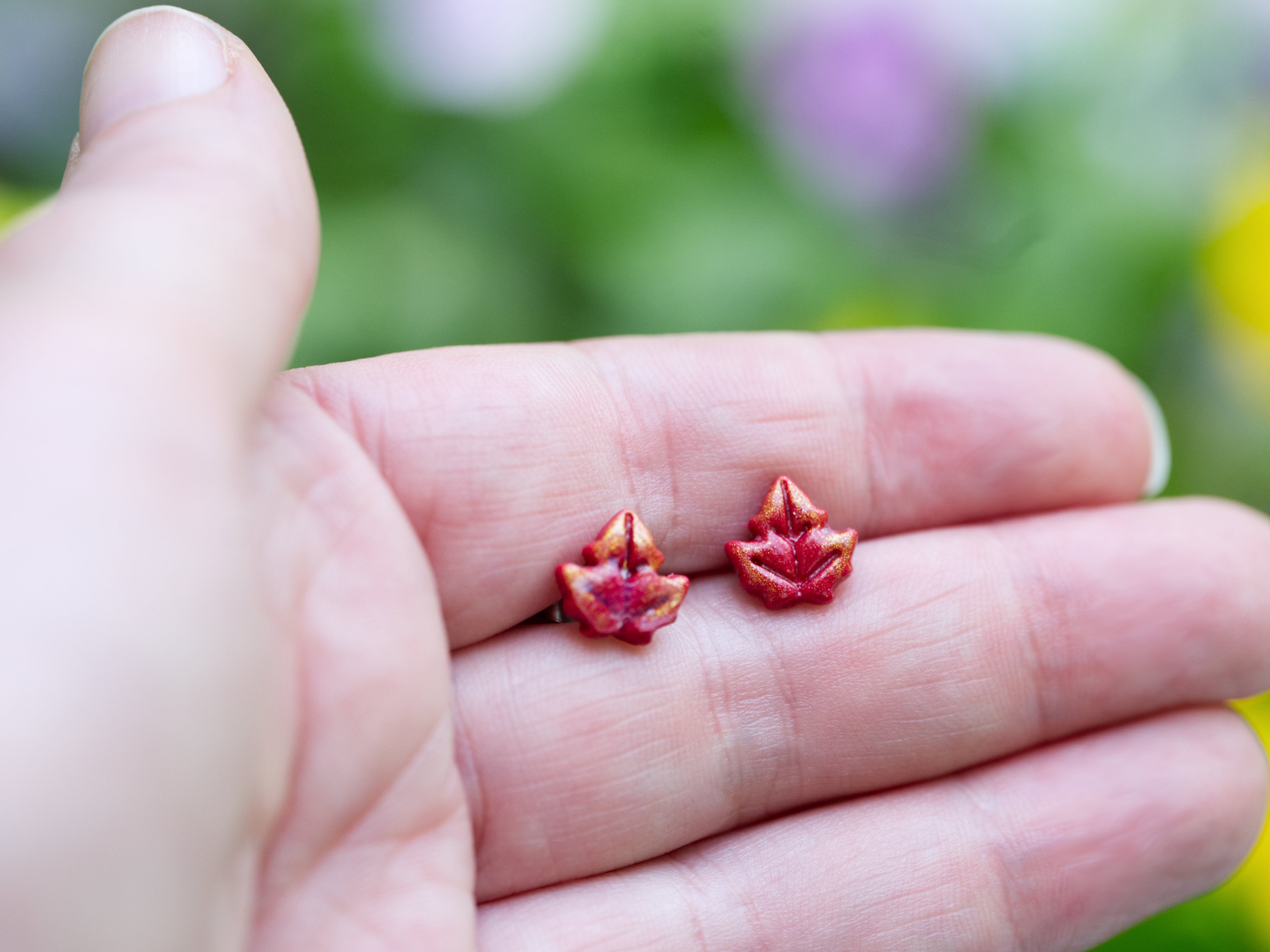 Red maple leaf polymer clay stud earrings shown at true size on Calithien Jewellery branded card held against purple flower garden background