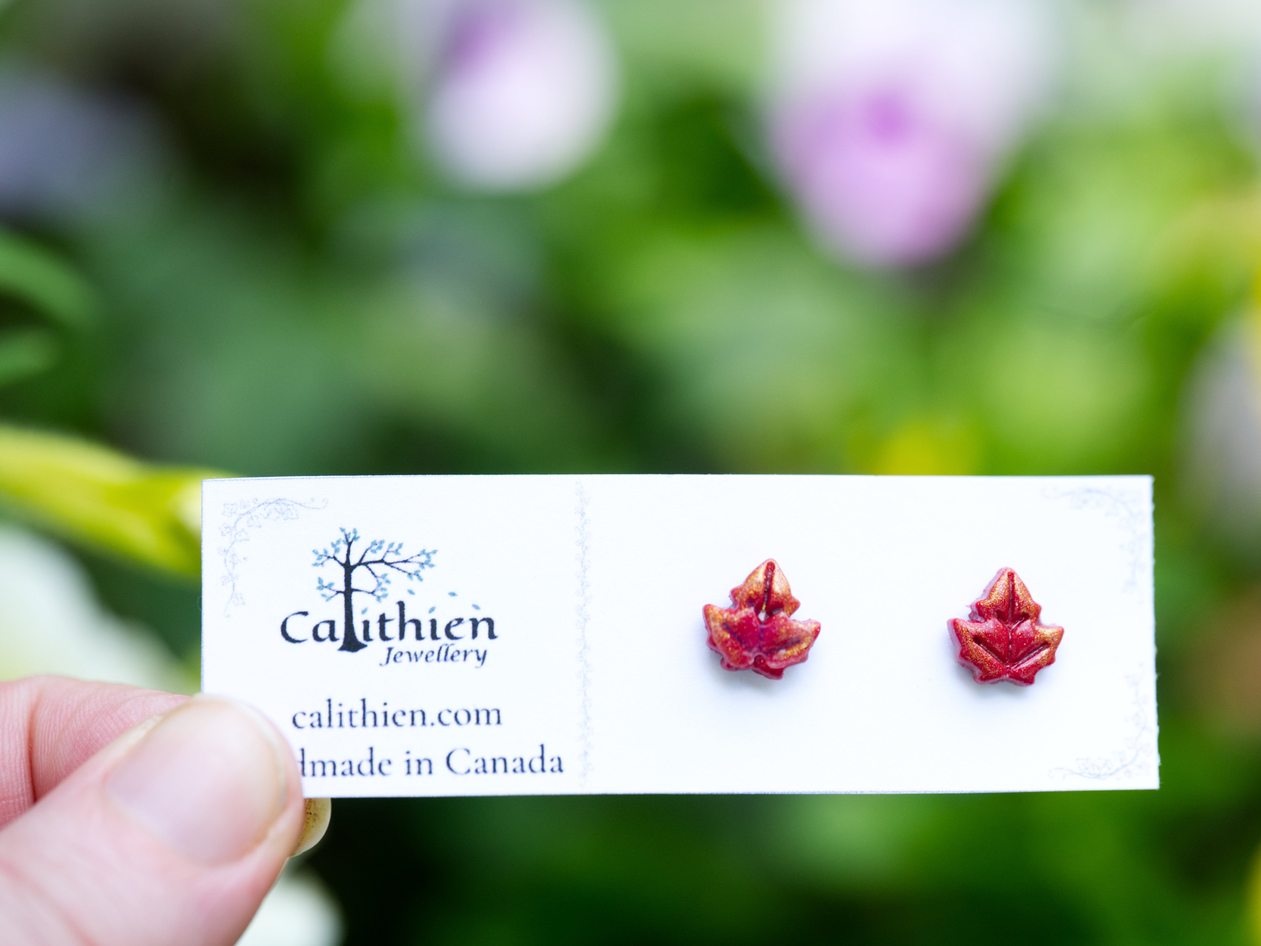 Red and gold shimmer handmade polymer clay maple leaf stud earrings held on card in hand against soft purple and green floral background