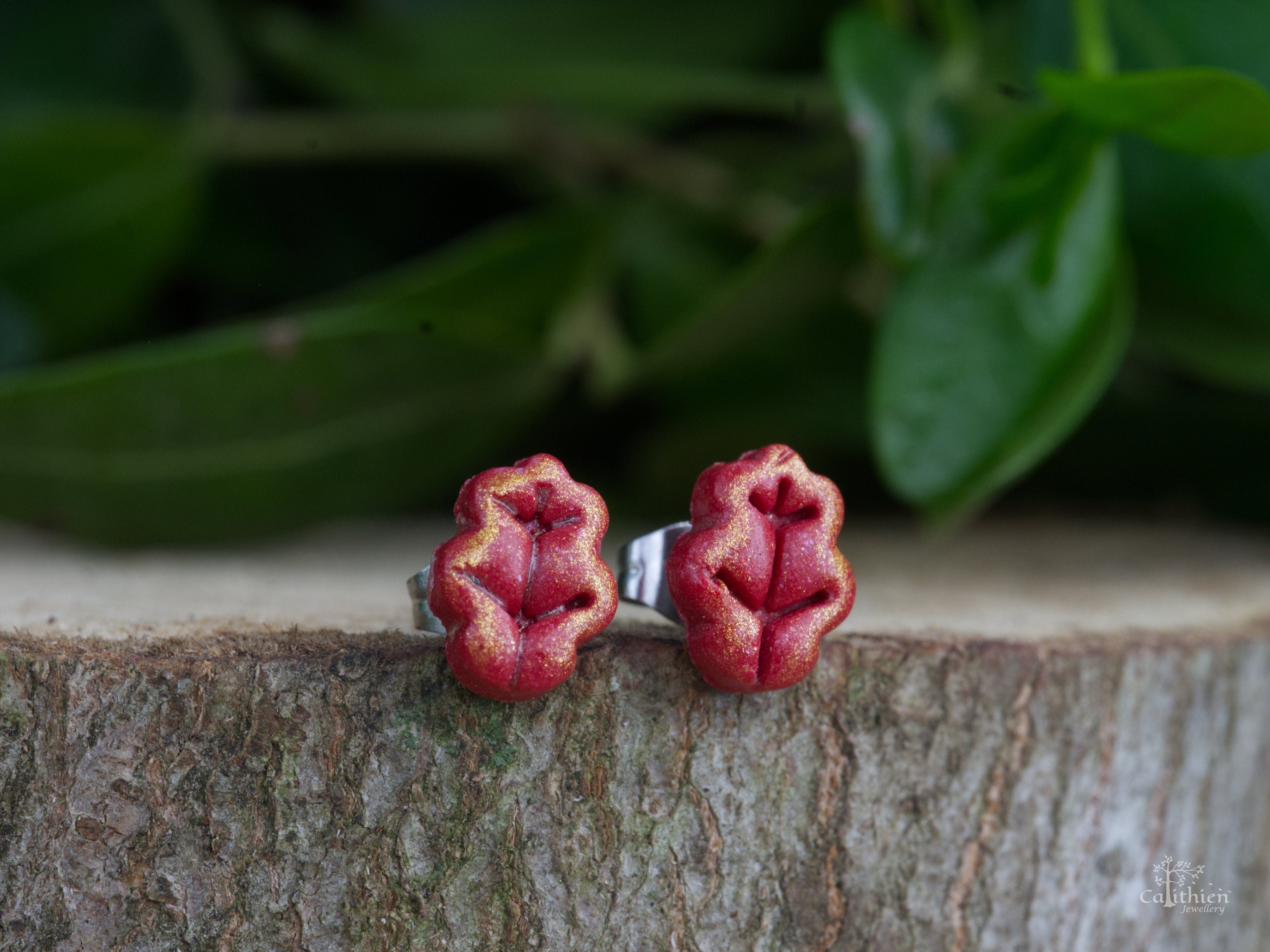 Handmade red polymer clay oak leaf stud earrings shown as a pair on natural bark surface with green garden foliage background