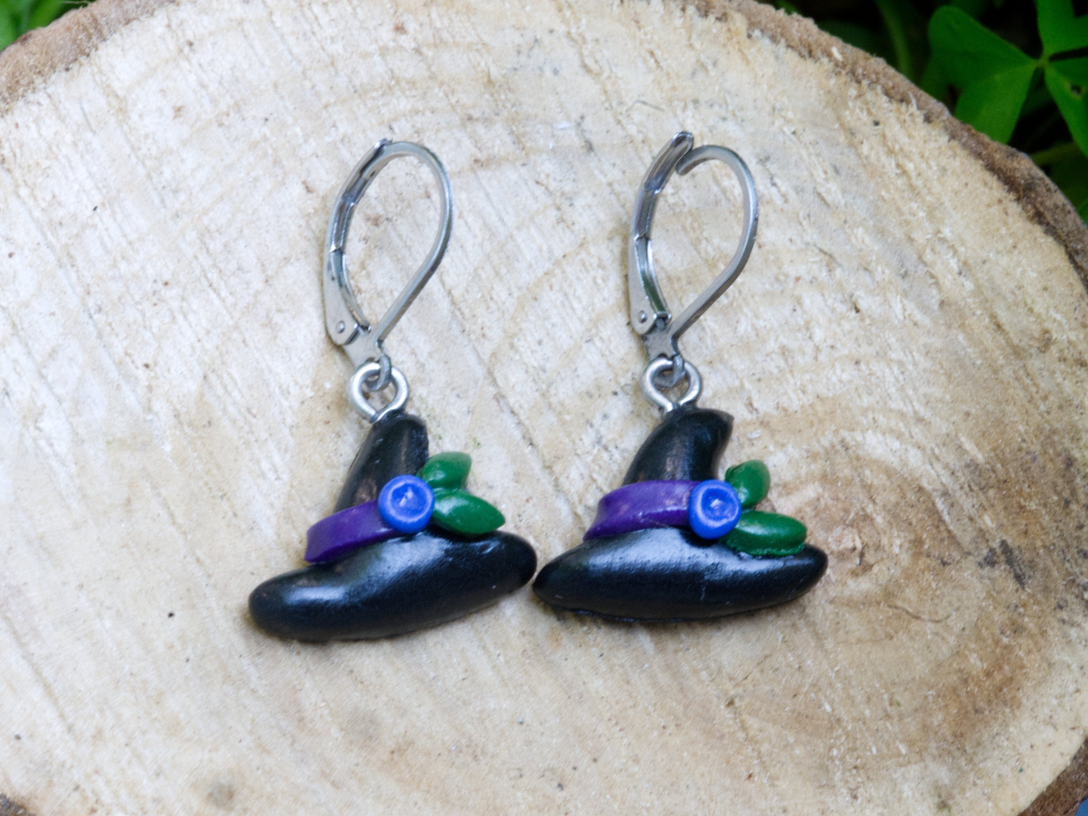 Handmade black polymer clay witch hat leverback earrings with purple ribbon blue flower and green leaf detail on natural wood surface