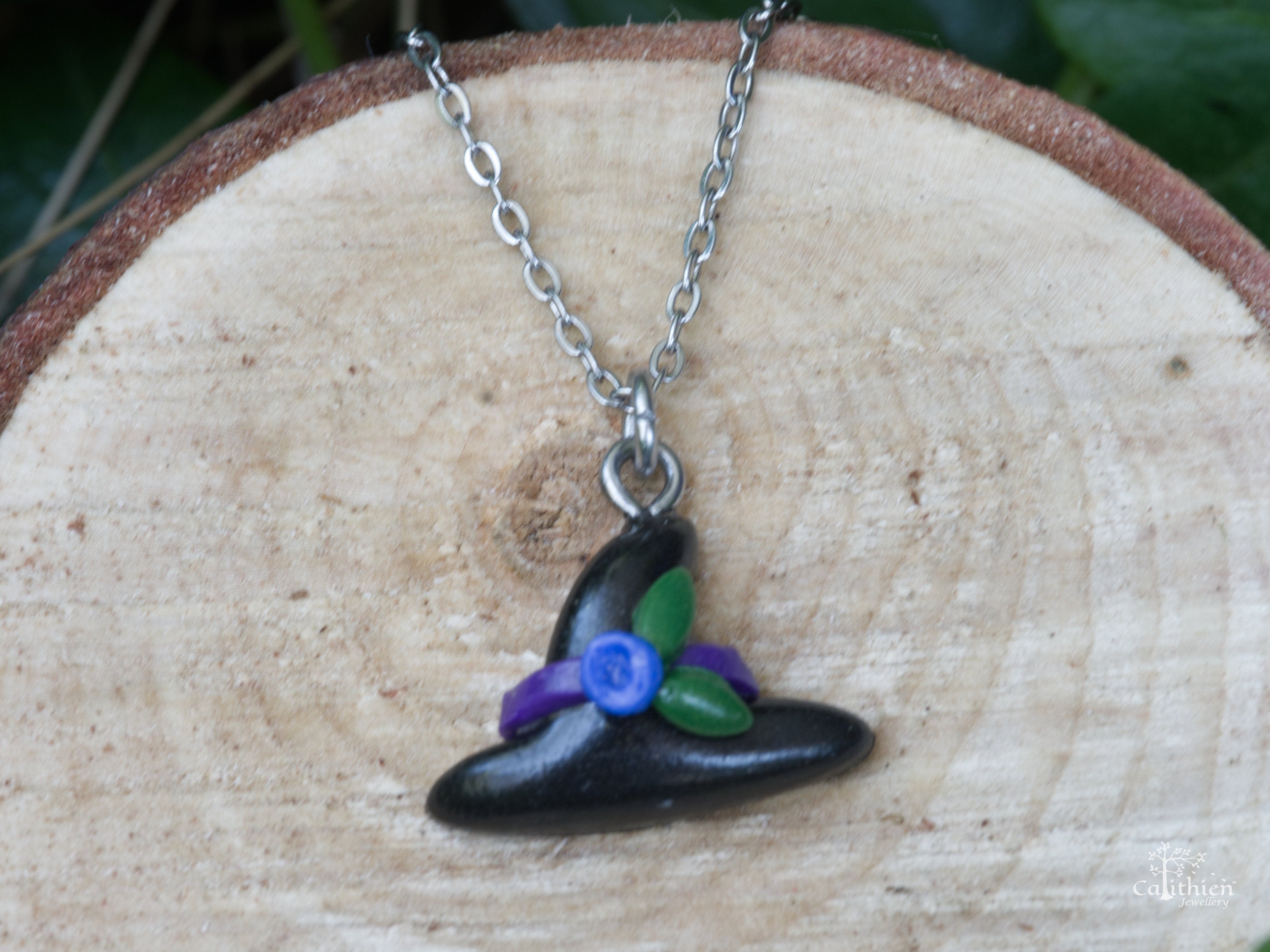 Handmade black polymer clay witch hat pendant necklace with purple ribbon blue flower and green leaf detail on natural wood surface