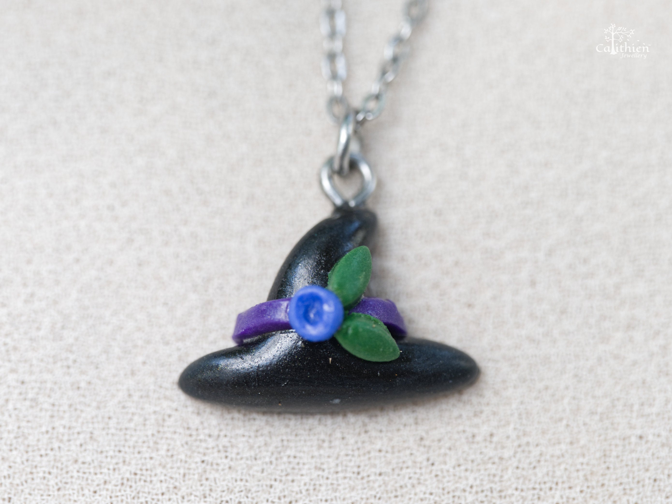 Close-up of black witch hat polymer clay pendant showing purple ribbon band, blue flower charm and green leaf accents with matte finish