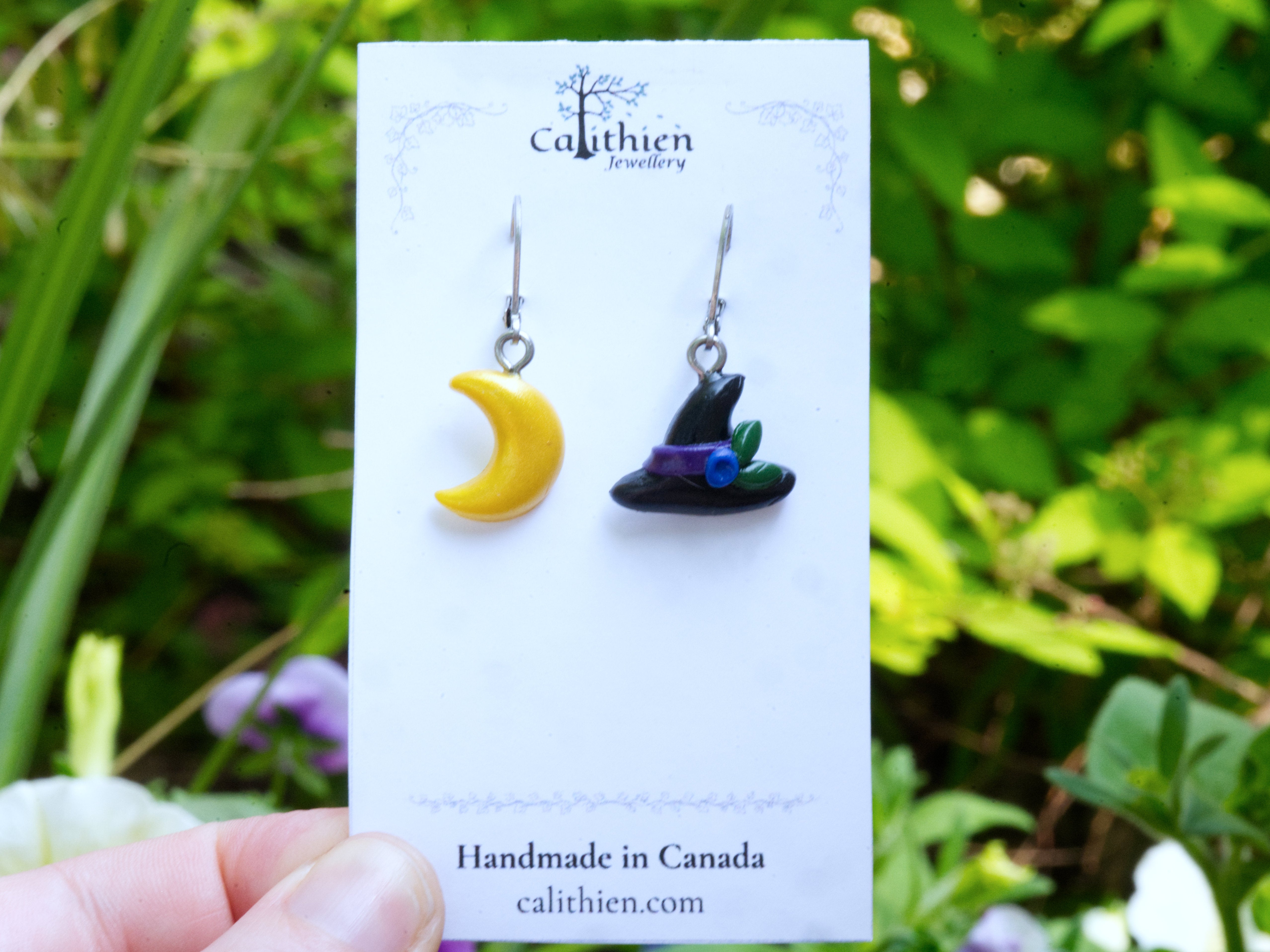 A pair of handmade polymer clay earrings on a white display card. The left earring is a yellow crescent moon, and the right earring is a black witch's hat with a purple and green band.