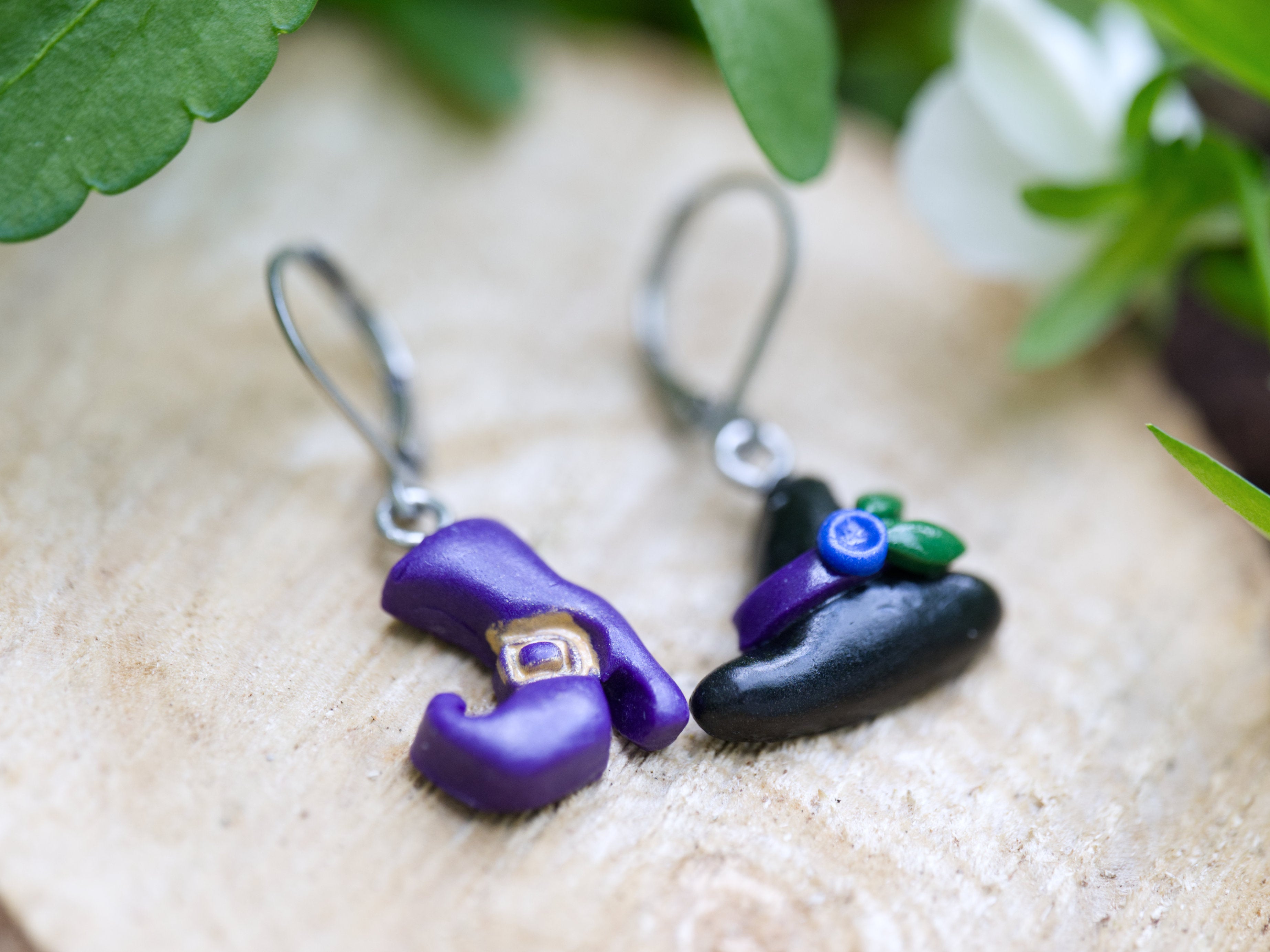 Close-up of mismatched witch hat earring with purple ribbon and blue flower beside purple polymer clay boot earring with gold buckle detail