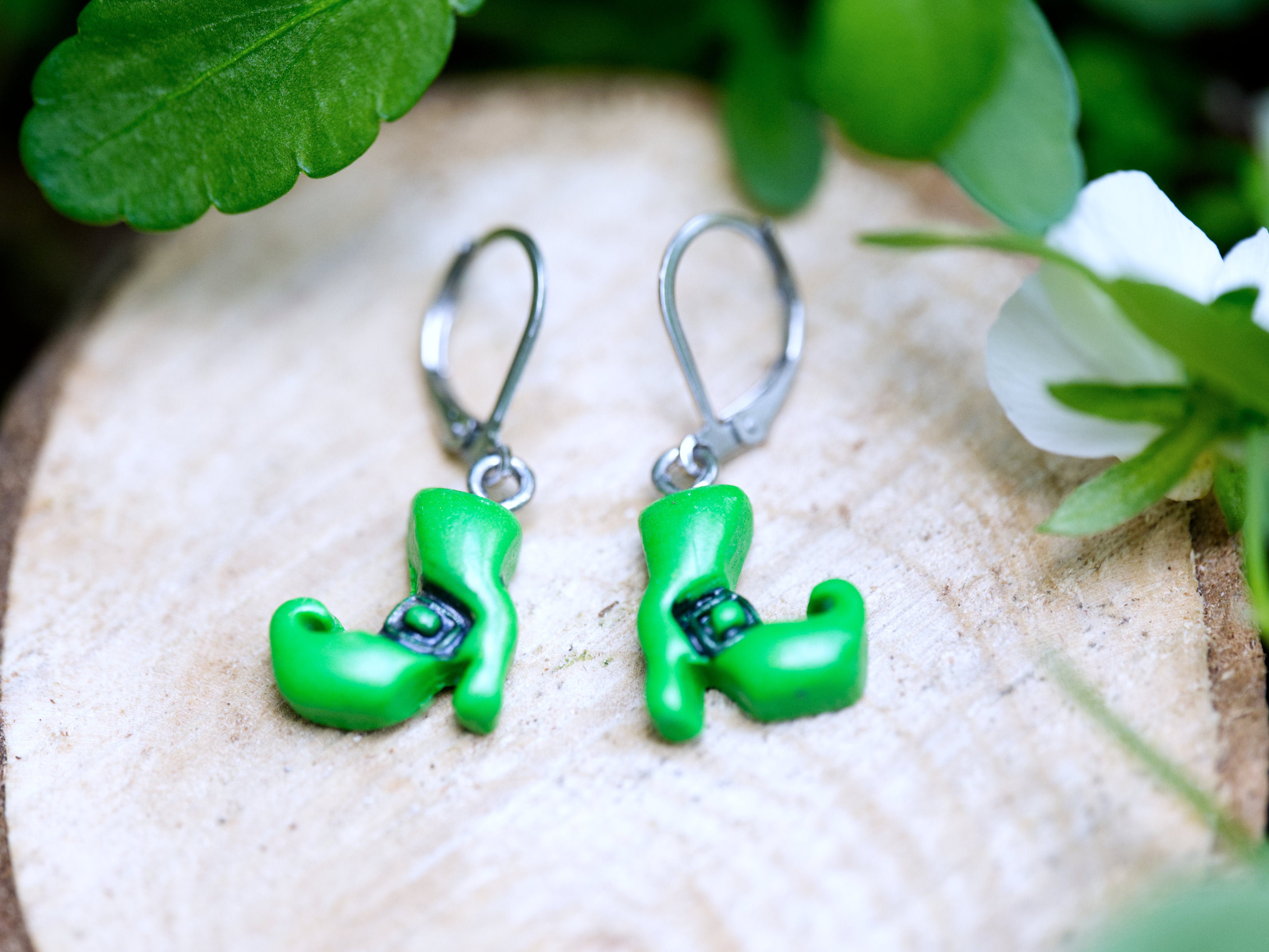 Handmade green polymer clay witch boot earrings on silver leverback hooks resting on natural wood slice with green succulent background