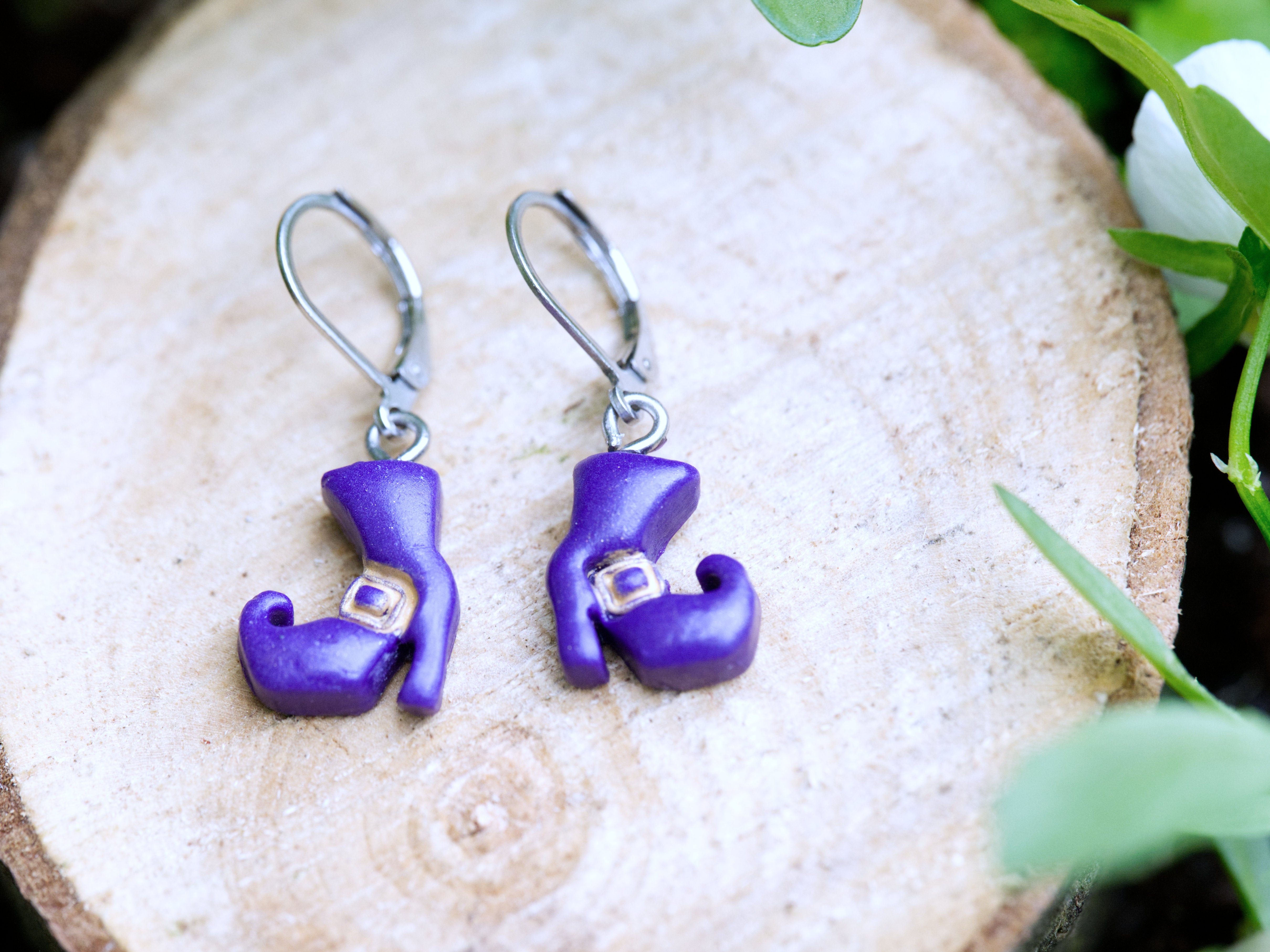Handmade purple polymer clay witch boot earrings on silver leverback hooks resting on natural wood slice with green floral background